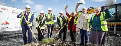 A group of people in safety vests and hard hats smiling and posing with shovels at a groundbreaking ceremony on a construction site.