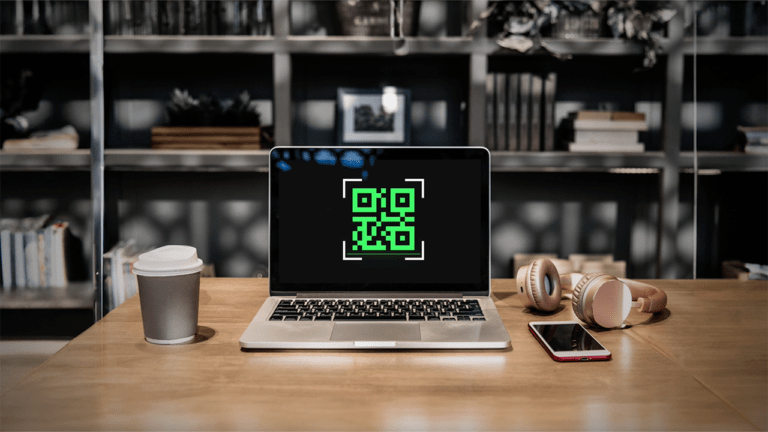 A laptop on a wooden desk displays a green QR code on its screen, with a coffee cup, headphones, and a smartphone beside it.